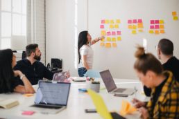 A woman with long black hair and glasses stands at the front of a room, pointing to a whiteboard full of colorful sticky notes. Her colleges look engaged in her workshop content.